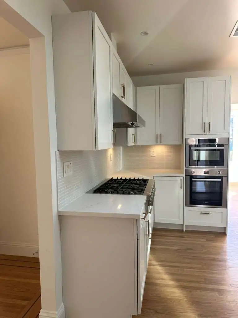 A modern kitchen with white cabinets, stainless steel appliances, a gas stove, and hardwood flooring. The space appears narrow with built-in ovens and under-cabinet lighting.