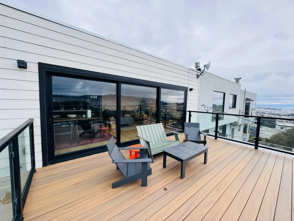 A modern rooftop deck with two chairs, a table, glass railing, and sliding doors leading into a house with a view of the city and hills in the background.