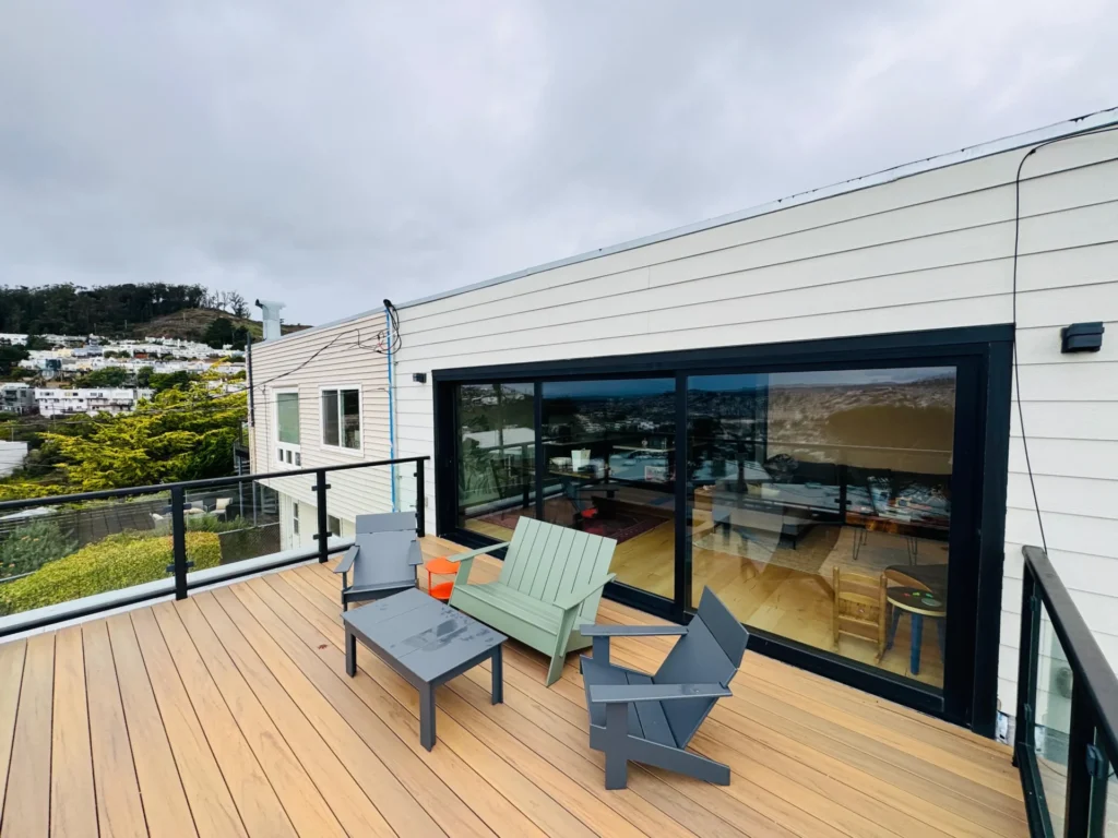 A rooftop deck with light wood flooring, three chairs, a bench, a small orange side table, and glass railing, next to a white building with large sliding glass doors.