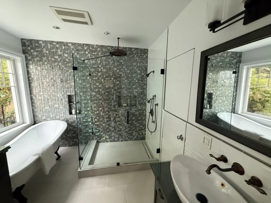 Modern bathroom with a glass shower, mosaic tile accent wall, clawfoot bathtub by a window, and a white sink with bronze fixtures.