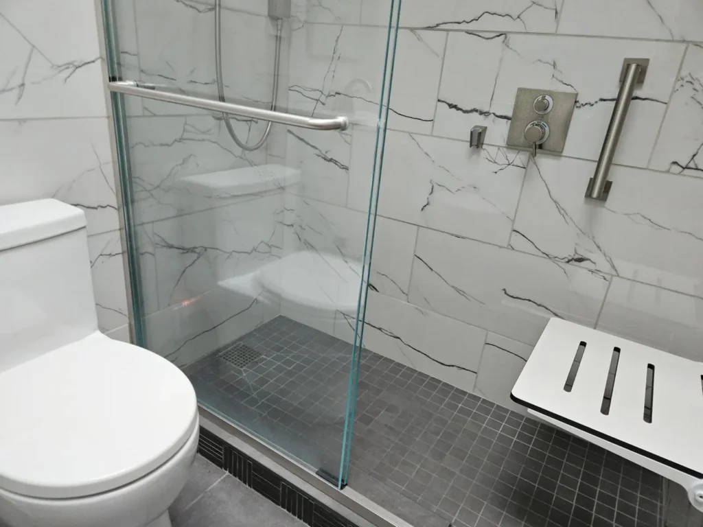 Accessible bathroom with a toilet, a glass-enclosed walk-in shower, grab bars, a wall-mounted shower, and a foldable shower bench on gray and white tile.