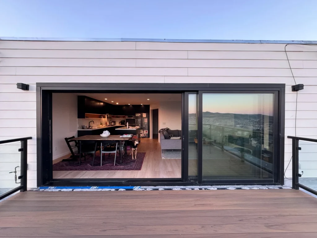 Siding Contractor. A modern home with large sliding glass doors opens to a deck, showing a kitchen, dining area, and living room with a view of mountains in the distance.