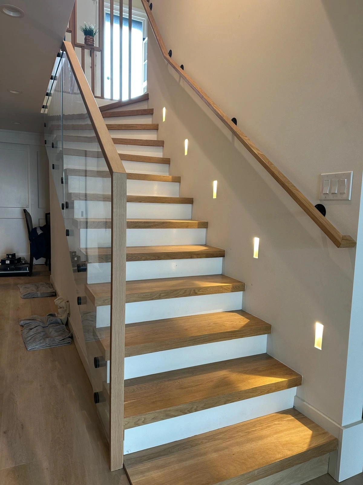 Home remodel. A modern wooden staircase with glass side panels, built-in wall lights, and a wooden handrail in a well-lit interior space.